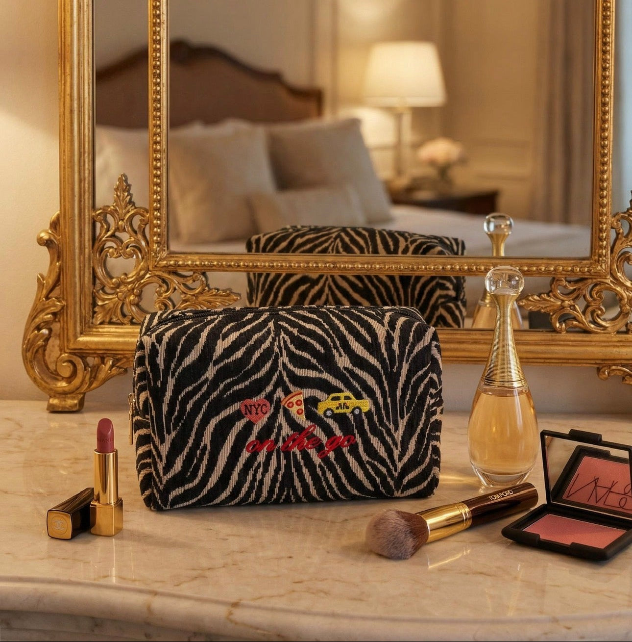 Makeup bag with zebra pattern and colorful text on a marble surface with makeup items, in front of a large ornate mirror.