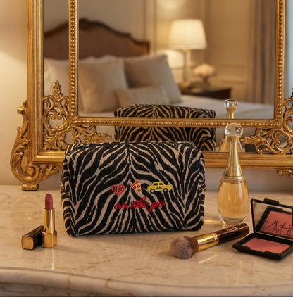 Makeup bag with zebra pattern and colorful text on a marble surface with makeup items, in front of a large ornate mirror.